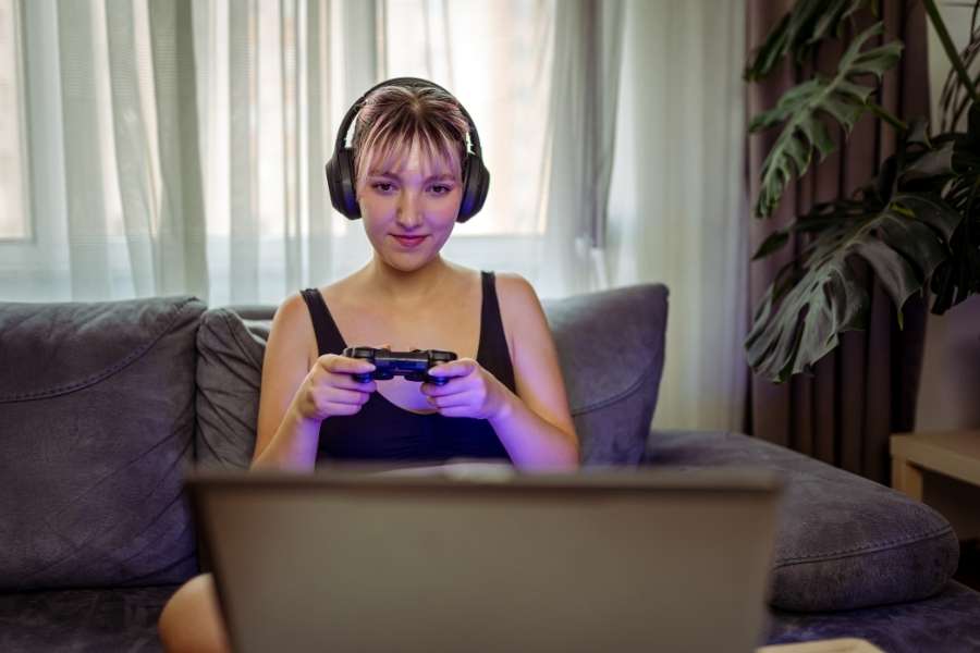 Une jeune femme joue avec une manette et un casque devant un laptop gaming posé sur ses genoux dans un salon.