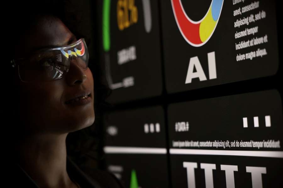 Profil d'une femme portant des lunettes transparentes qui regarde un grand écran affichant des données informatiques et le logo AI.