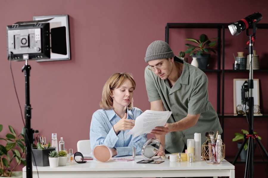 Une femme assise à un bureau de maquillage et un homme debout relisent un script imprimé sur un plateau de tournage vidéo.